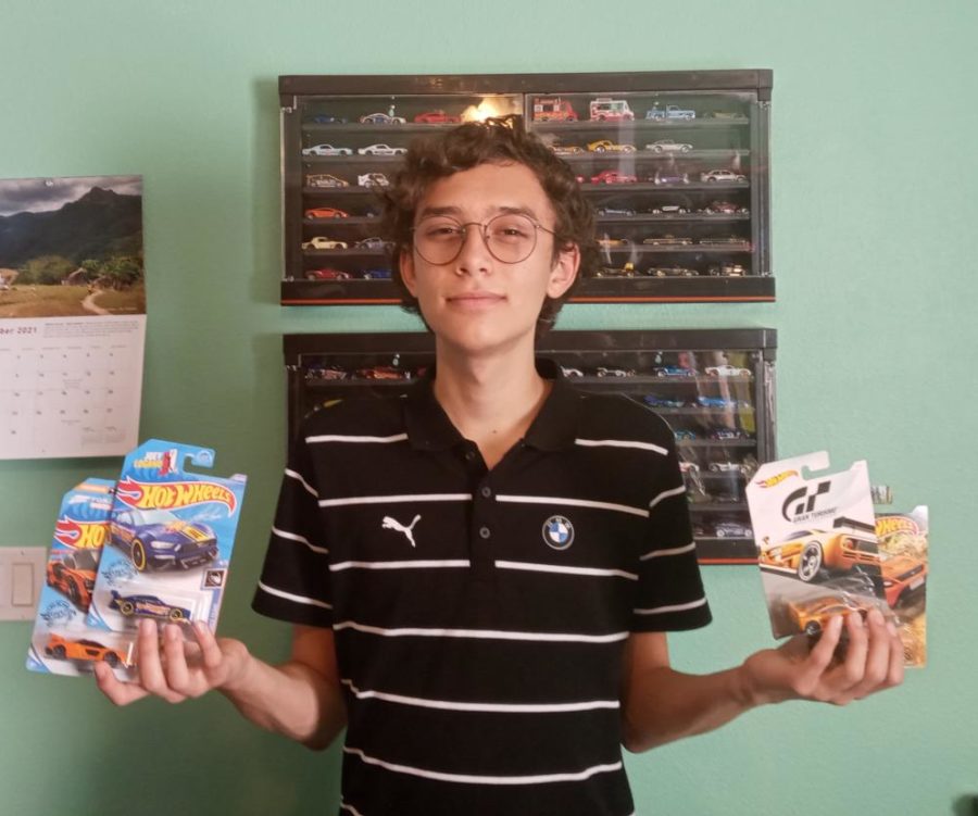 Julian Reyes-Rogachevsky holds up 4 of his newer boxed hot wheels standing in front of his showcases containing the majority of his most prized cars. 